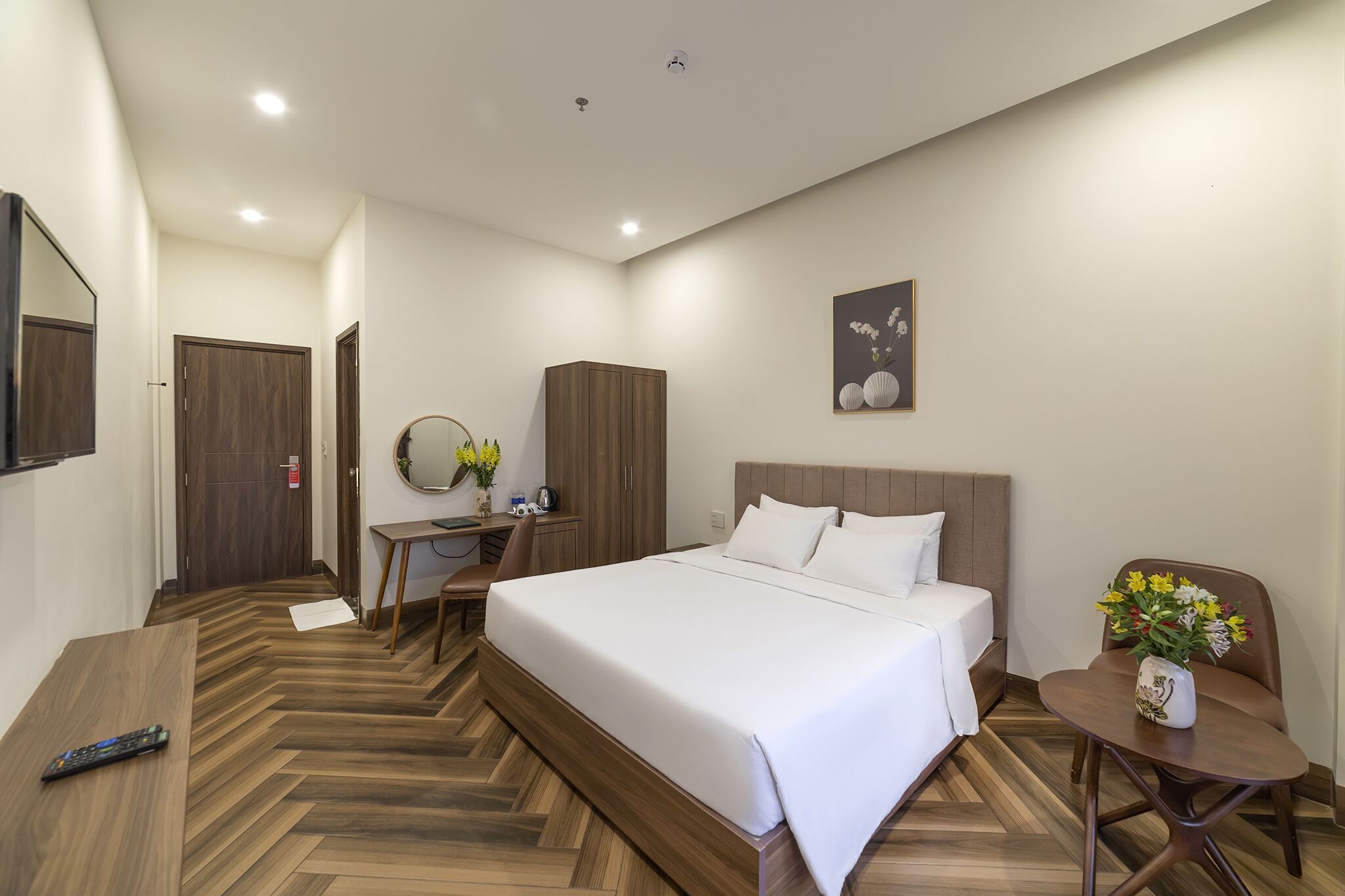 A hotel room with a large bed, a desk with a chair, a small table with a vase of flowers, and a mirror on the wall.