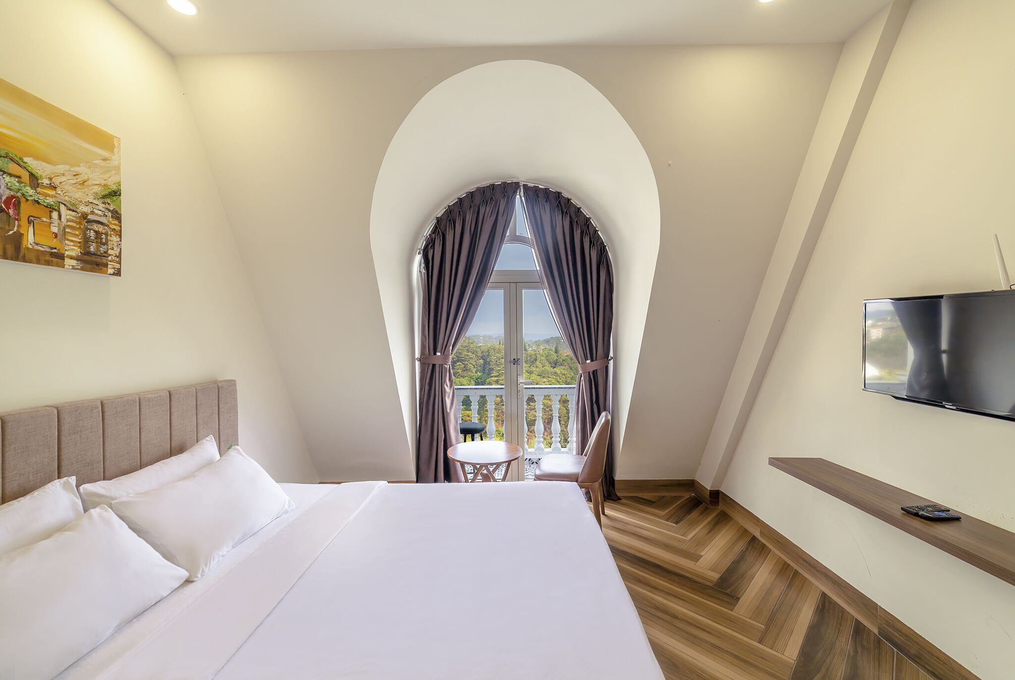 Une chambre d’hôtel avec un grand lit, un téléviseur fixé au mur, un balcon avec une table et des chaises, et un tableau accroché au mur.