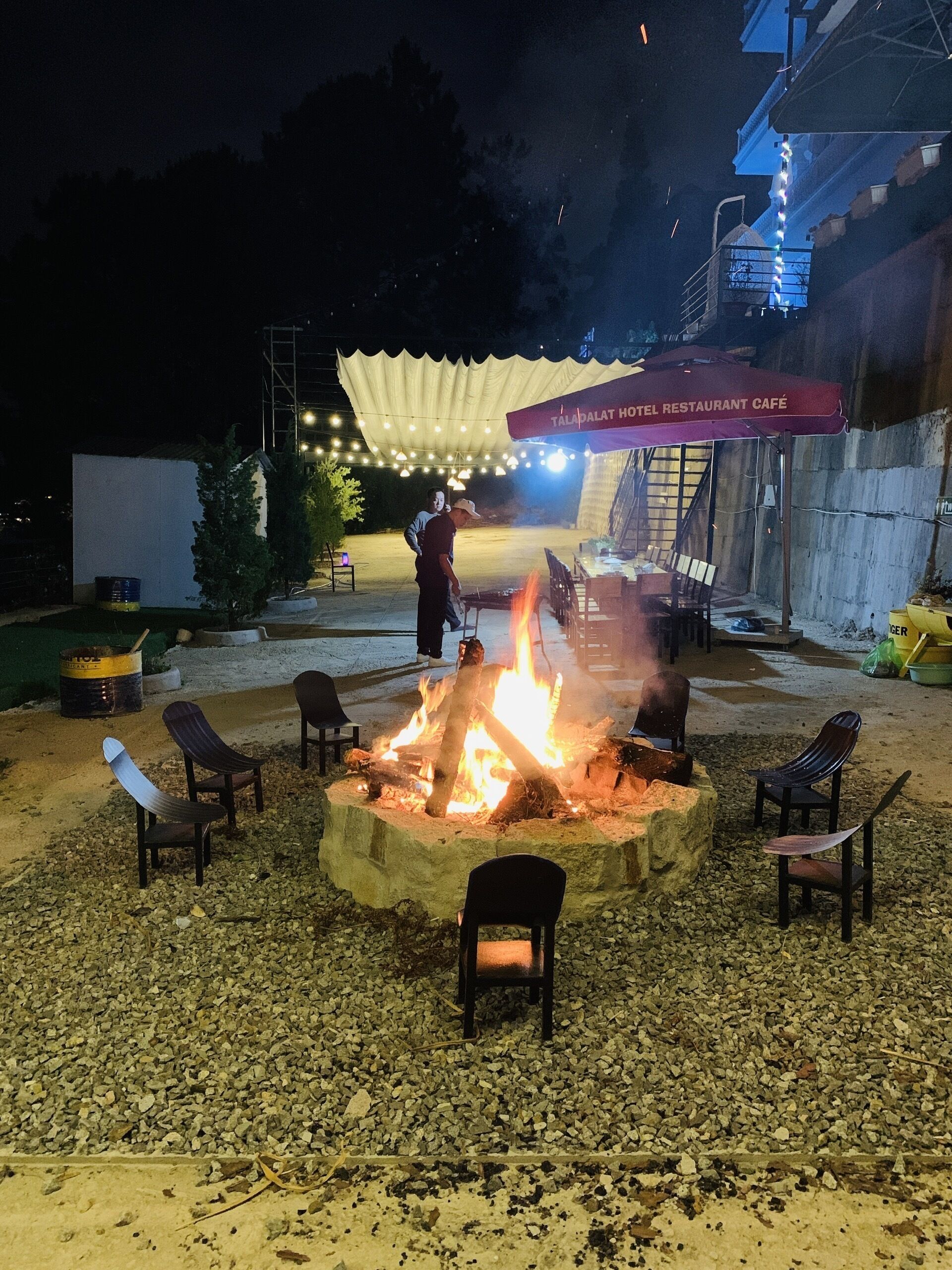 An outdoor seating area with a fire pit surrounded by chairs, under a marquee with string lights.