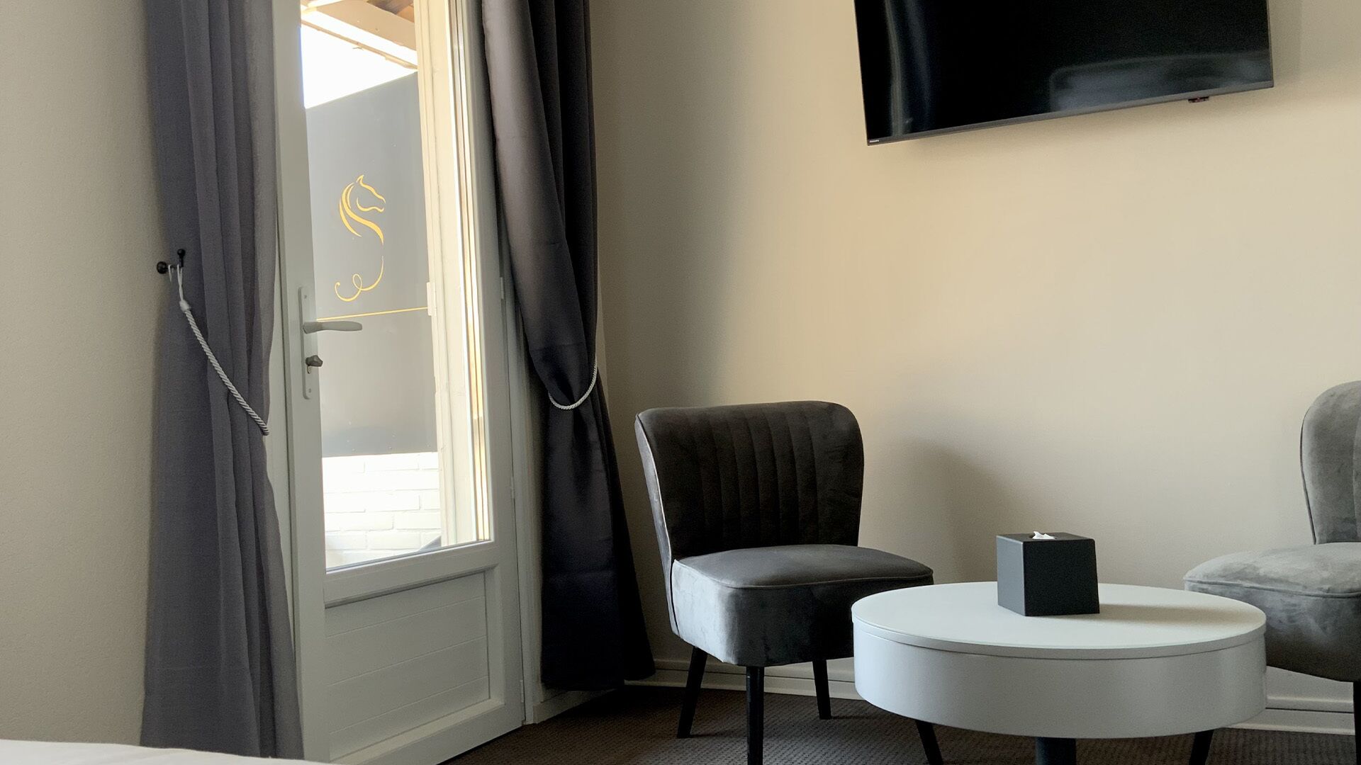 Een hotelkamer met een bed, een ronde witte tafel, twee fauteuils, een aan de muur bevestigde televisie en een raam met gordijnen.