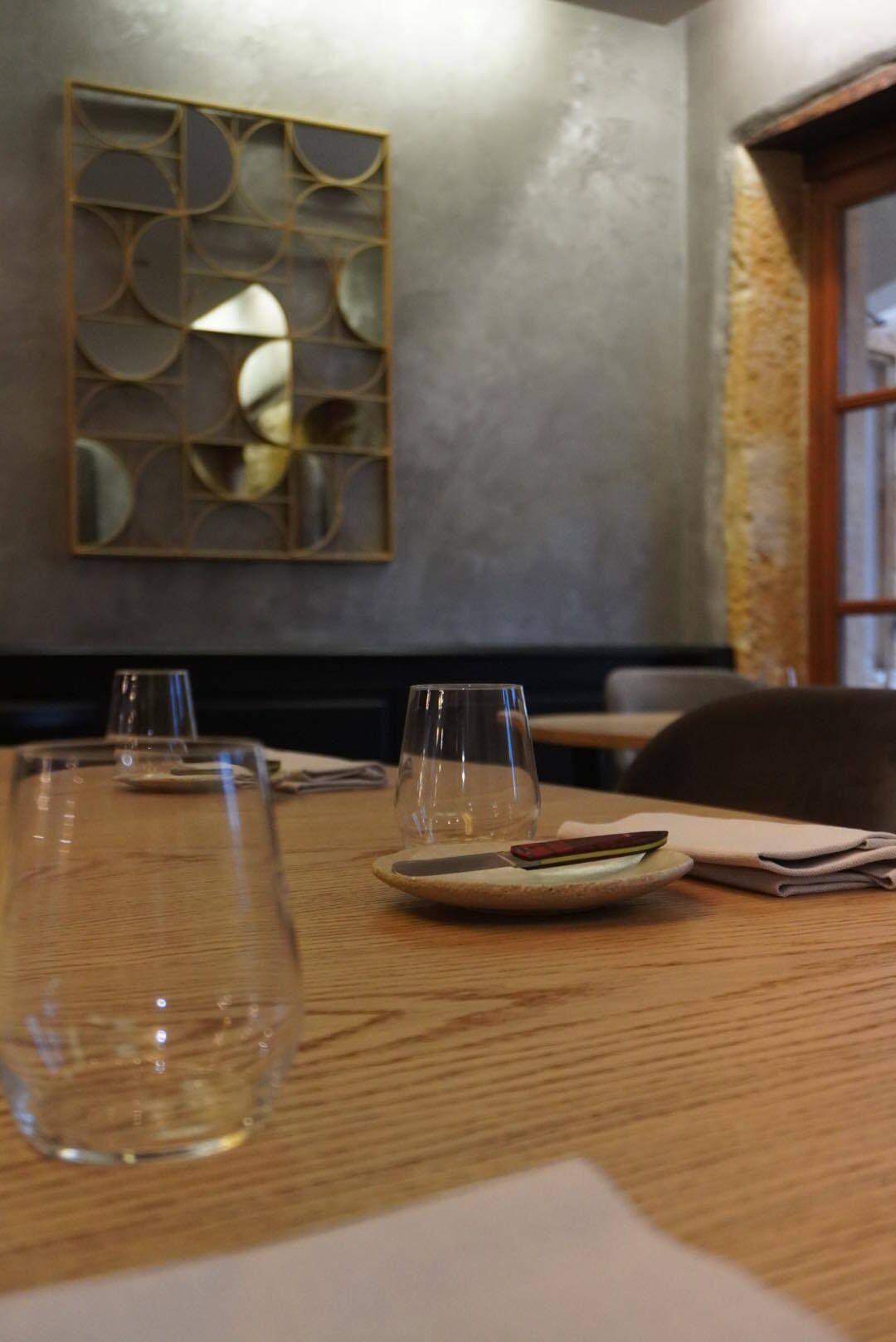 A dining table with two empty wine glasses, a plate with a napkin, and a menu. A decorative wall panel is visible in the background.