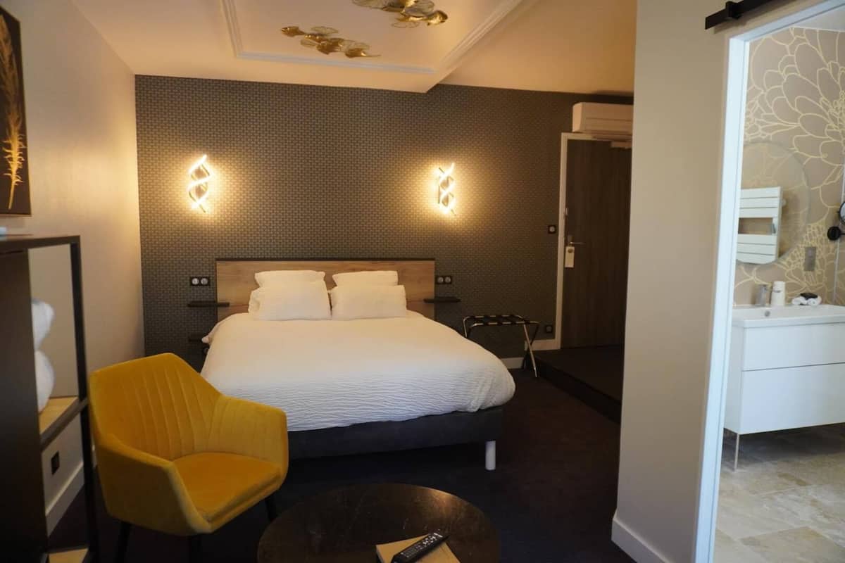 A hotel room with a large bed, a yellow chair, a small round table, and a white bedside table with a remote control.