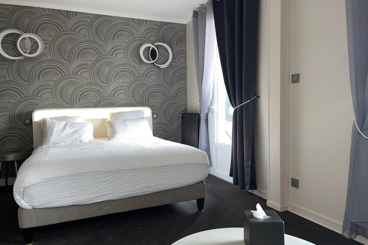 A modern hotel room with a large bed, a round bedside table, a bedside lamp, and a window with curtains.
