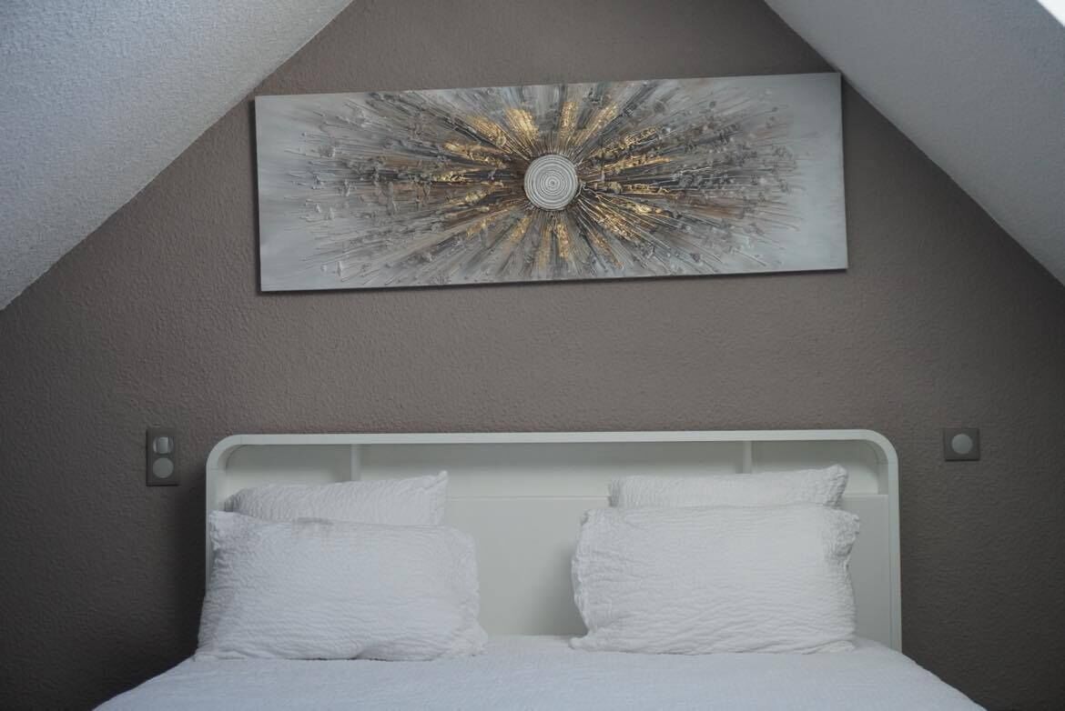 Une chambre avec un lit blanc, un mur gris et un plafond sous un toit en pente.