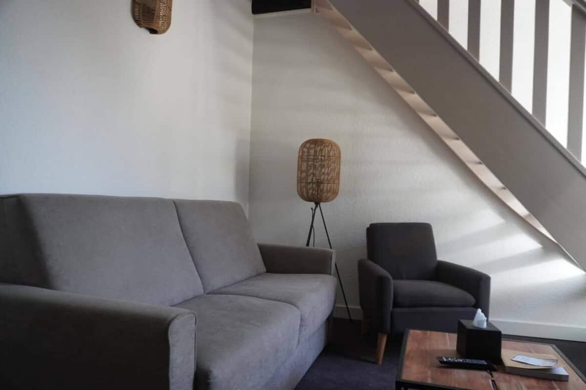 A modern living room with a grey sofa, a black armchair, and a wooden coffee table. There is a floor lamp with a woven base and a small table lamp on the coffee table. A staircase with a wooden railing is visible in the background.