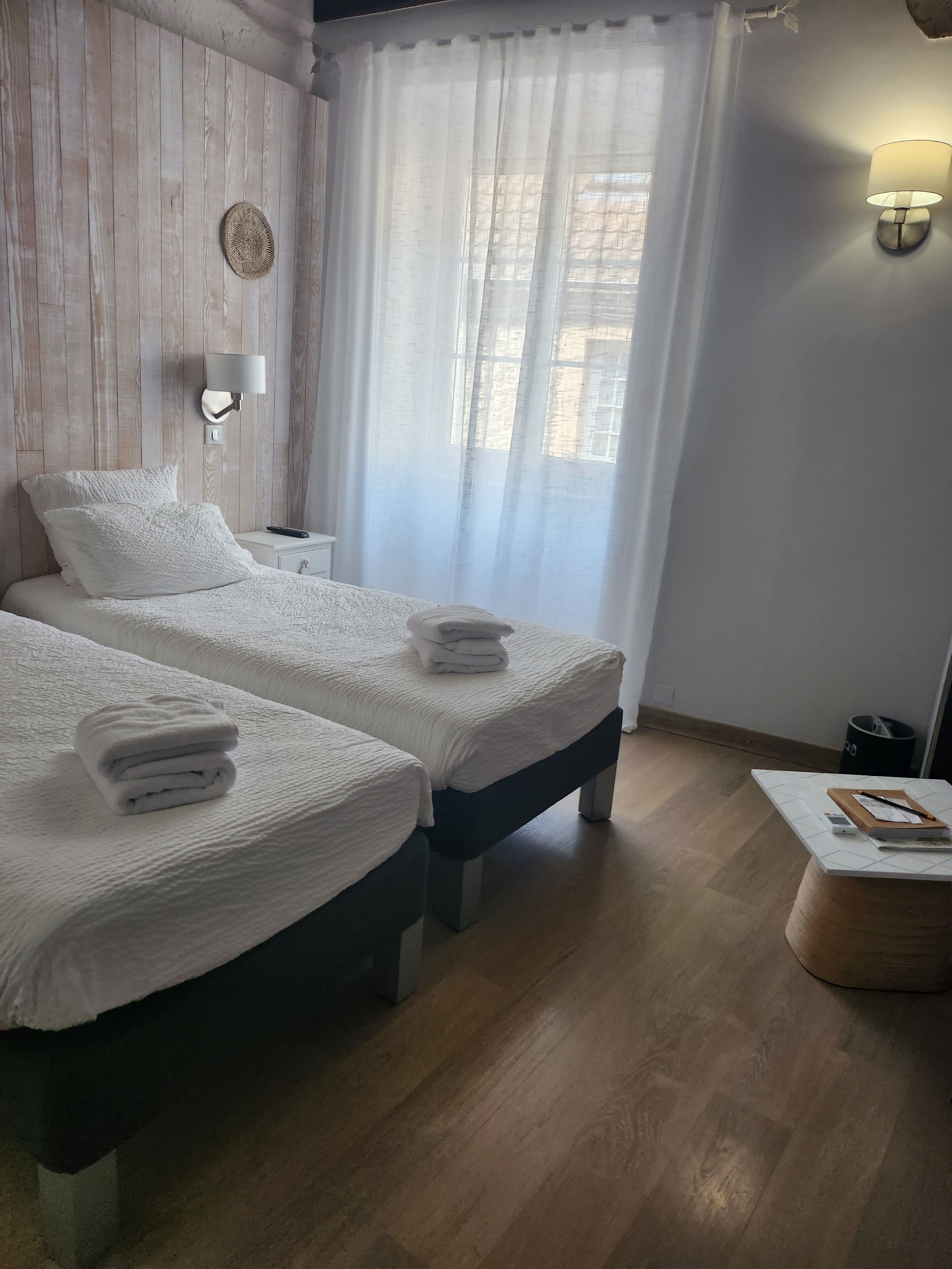 Et hotelværelse med to senge, et natbord, en lampe, et lille bord med en notesbog og en skraldespand.