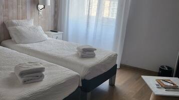 Et hotelværelse med to senge, et natbord, en lampe, et lille bord med en notesbog og en skraldespand.