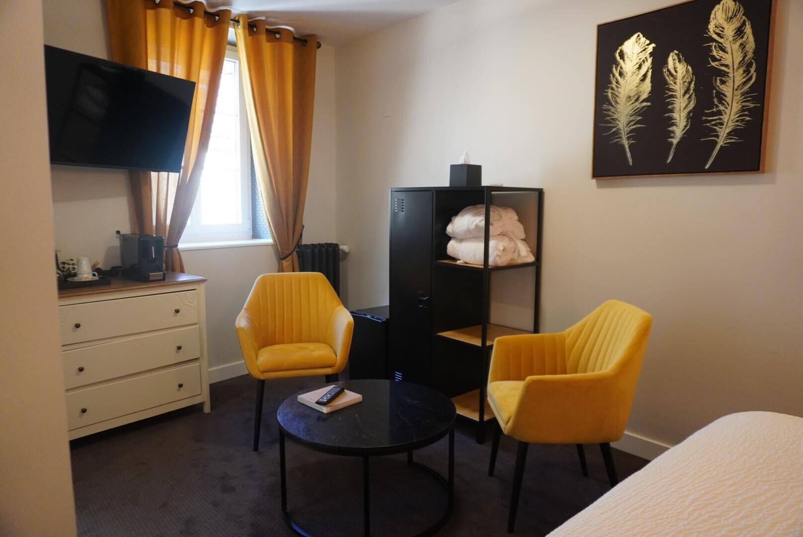 Quarto de hotel com duas cadeiras amarelas, uma mesa preta, uma televisão, um armário com toalhas dobradas e um quadro com penas na parede.