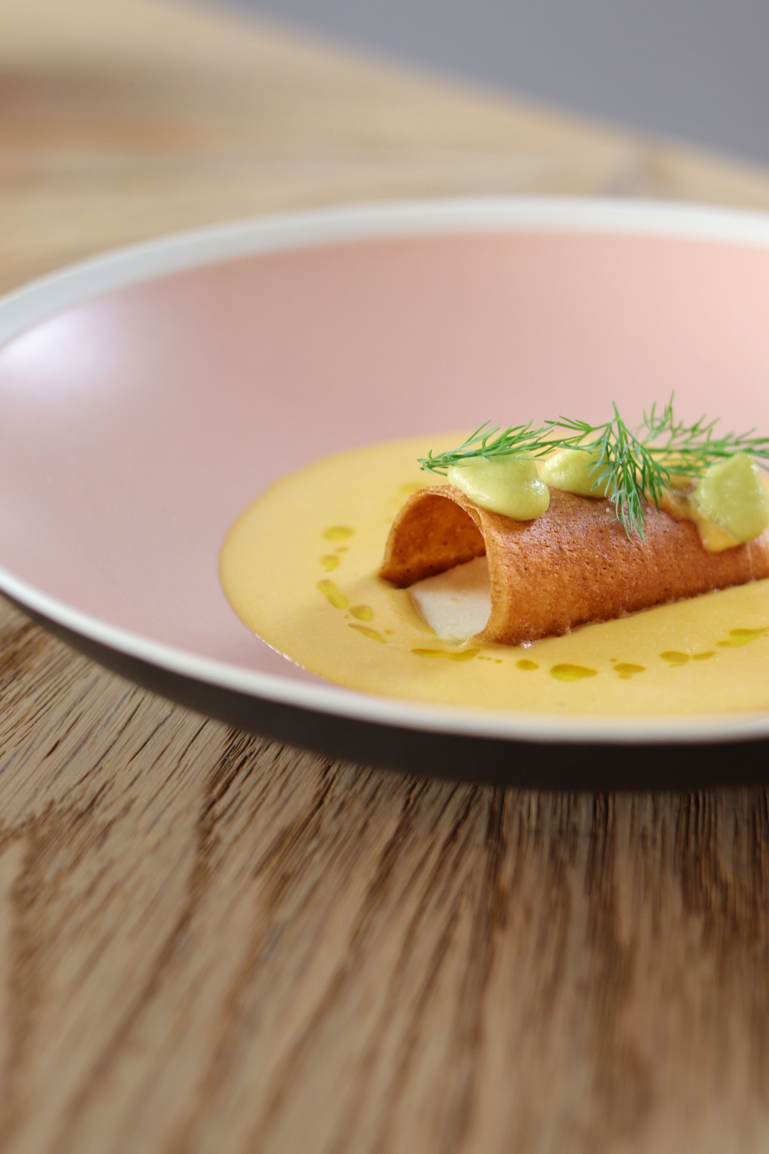 A plate with a rolled item, possibly a pastry, garnished with a green herb and a yellow sauce.
