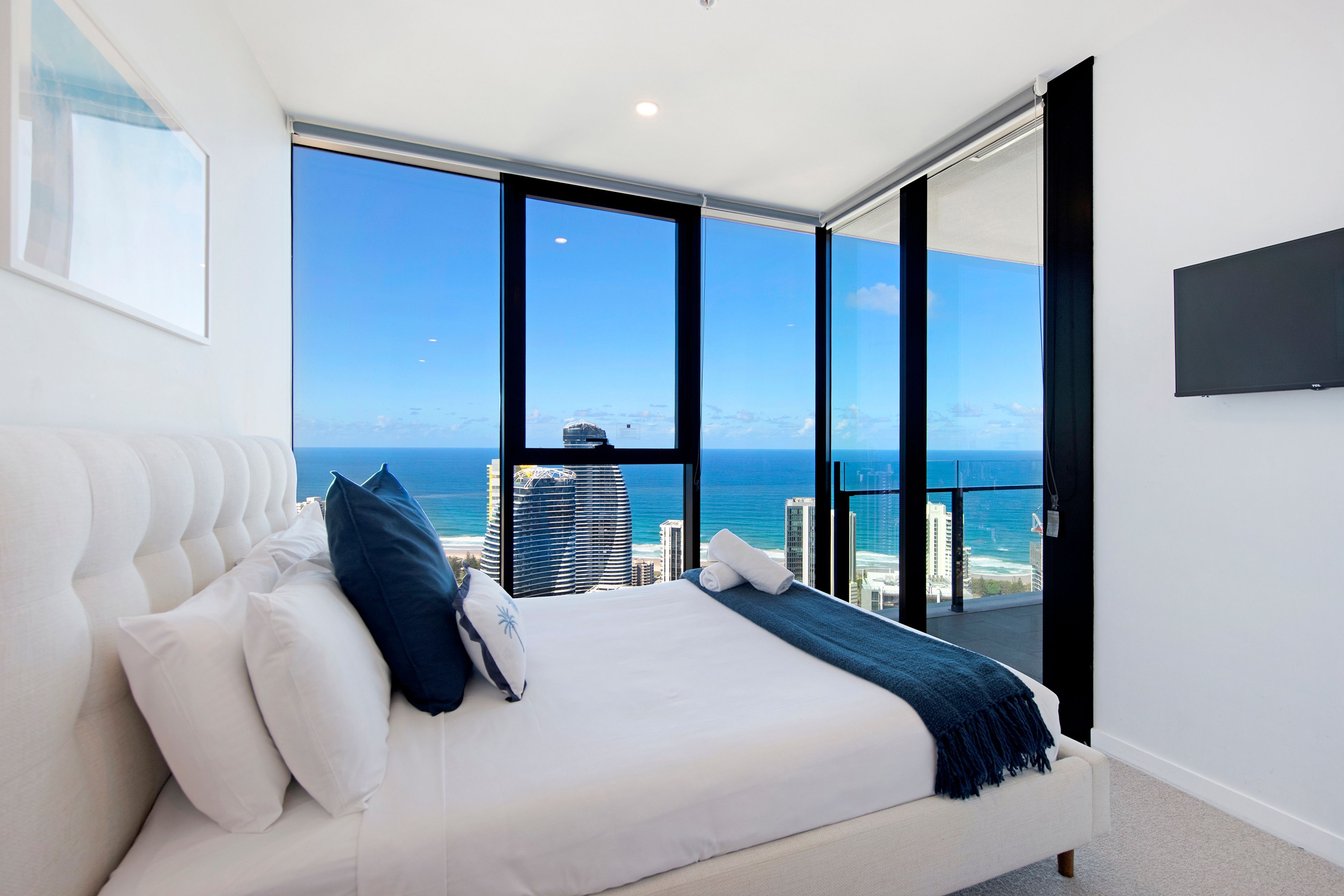 A bedroom with a large bed, white bedding, and a view of the ocean and buildings.