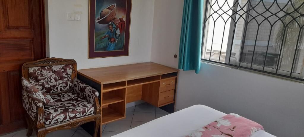 Une pièce avec un bureau en bois, un fauteuil à motifs, un lit recouvert d’une couvre-lit à fleurs, un tableau accroché au mur et une fenêtre ornée d’une grille en métal.