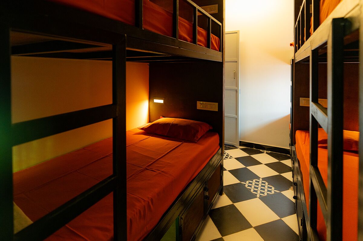 A dormitory room with bunk beds, a checkered floor, and a single bed with an orange blanket.