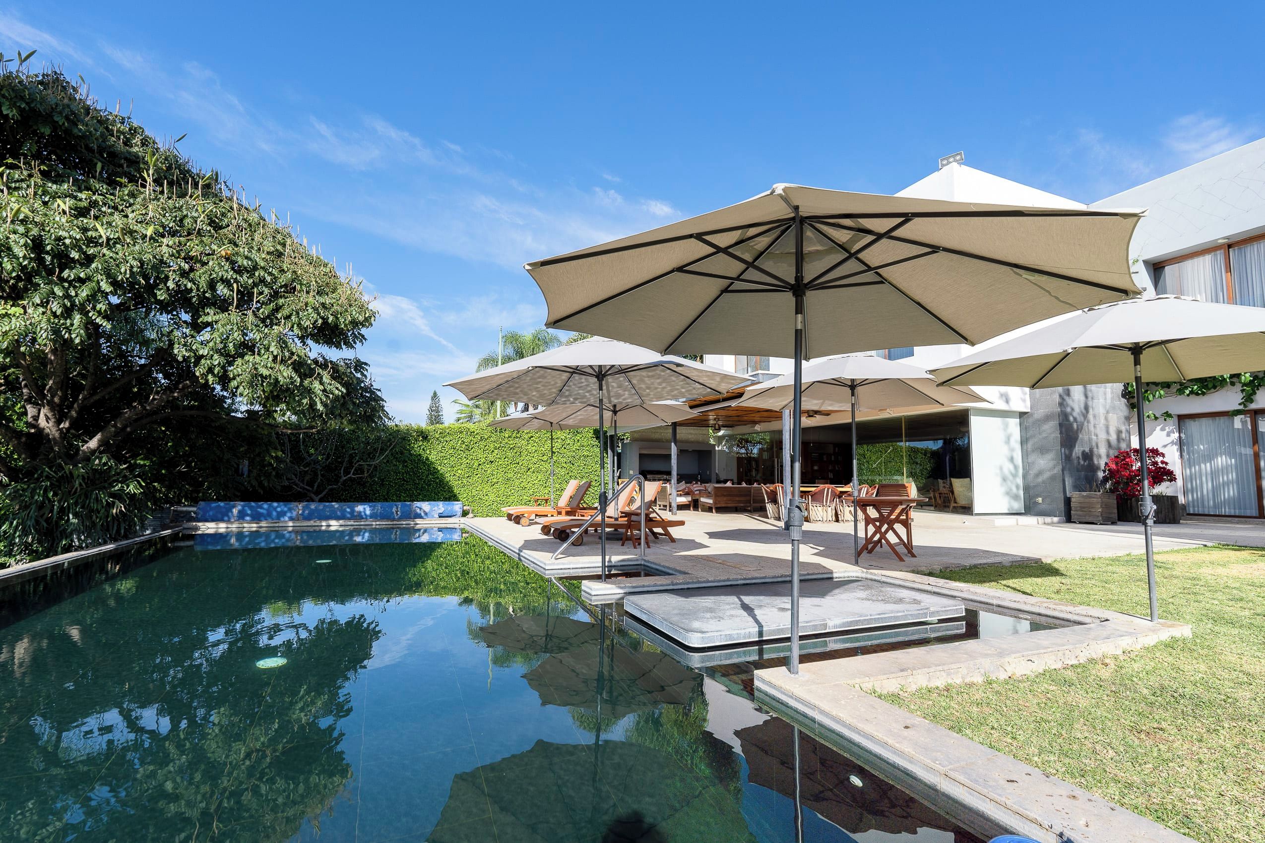 A swimming pool with lounge chairs and umbrellas, surrounded by greenery and a modern building.