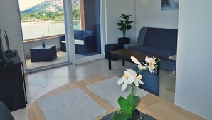 A modern living room with a sofa, a coffee table, and a potted plant. There is a sliding glass door leading to a balcony with a view of a mountain and a body of water. - Plava Laguna - Direkt am Strand und mit Meerblick (Seline)