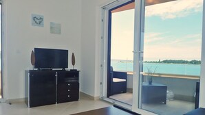A modern living room with a flat-screen TV, a dark wooden cabinet, and a large sliding glass door leading to a balcony with a view of a body of water. - Plava Laguna - Direkt am Strand und mit Meerblick (Seline)