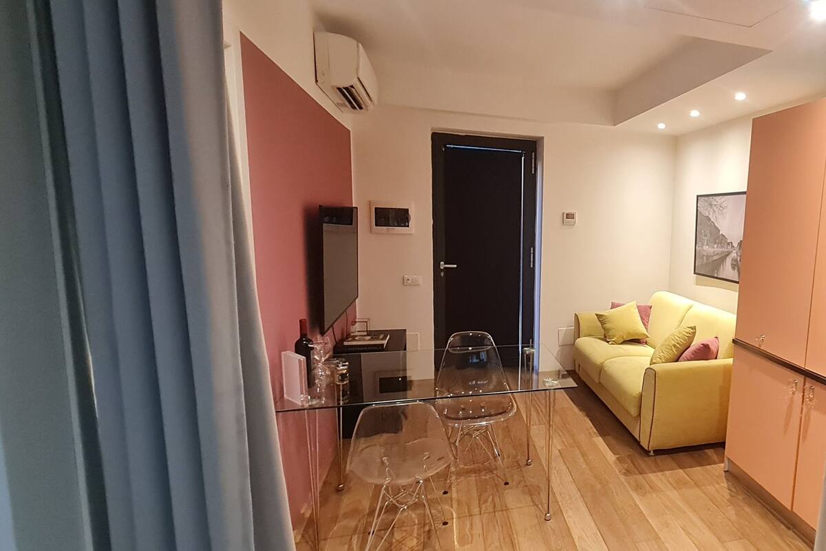 Ein Wohnbereich mit Glastisch, zwei Stühlen, einem gelben Sofa mit Kissen, einem Kleiderschrank und einer Tür.