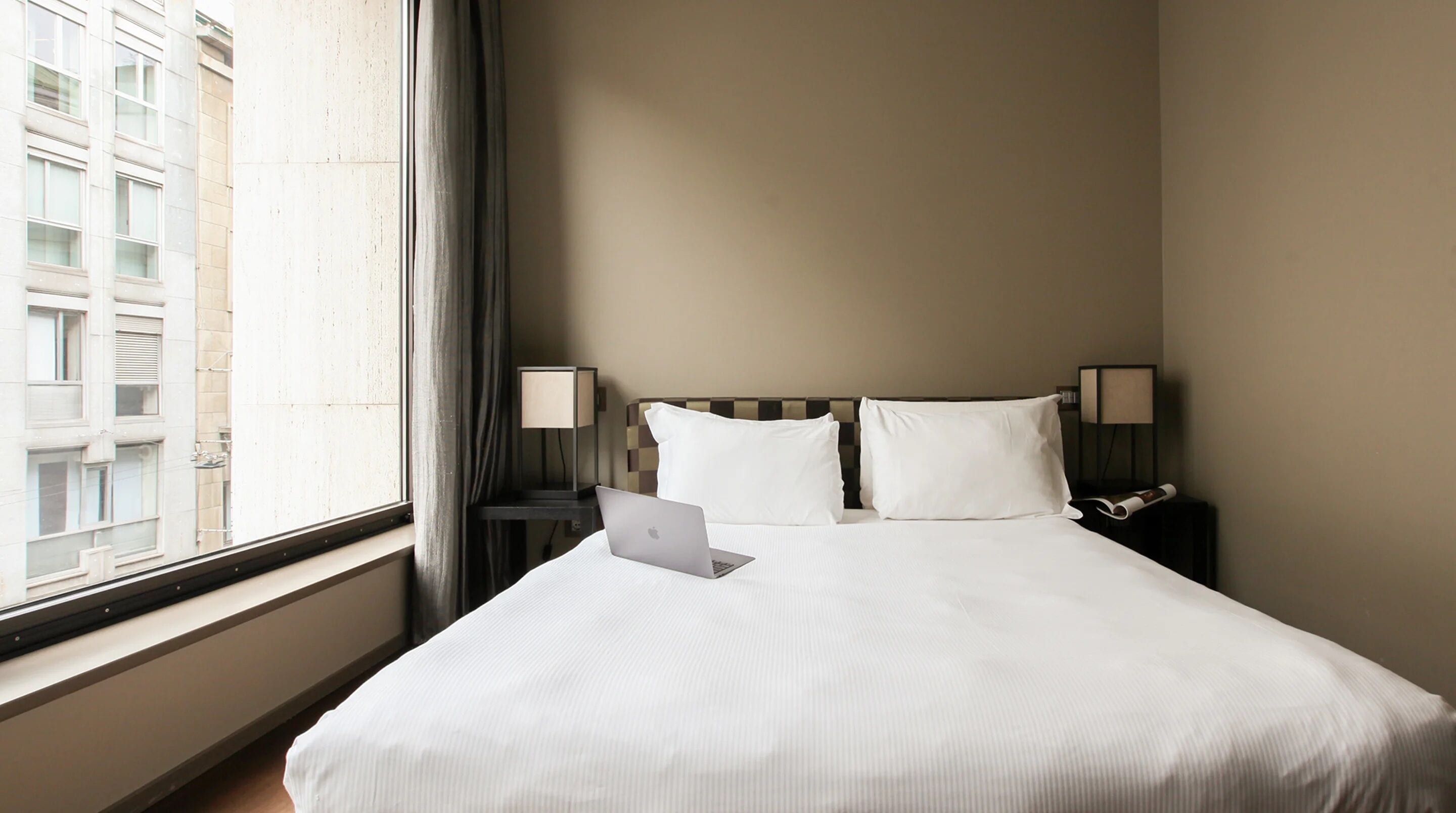A hotel room with a large bed, two bedside lamps, a laptop on the bed, and a window with a view of buildings.