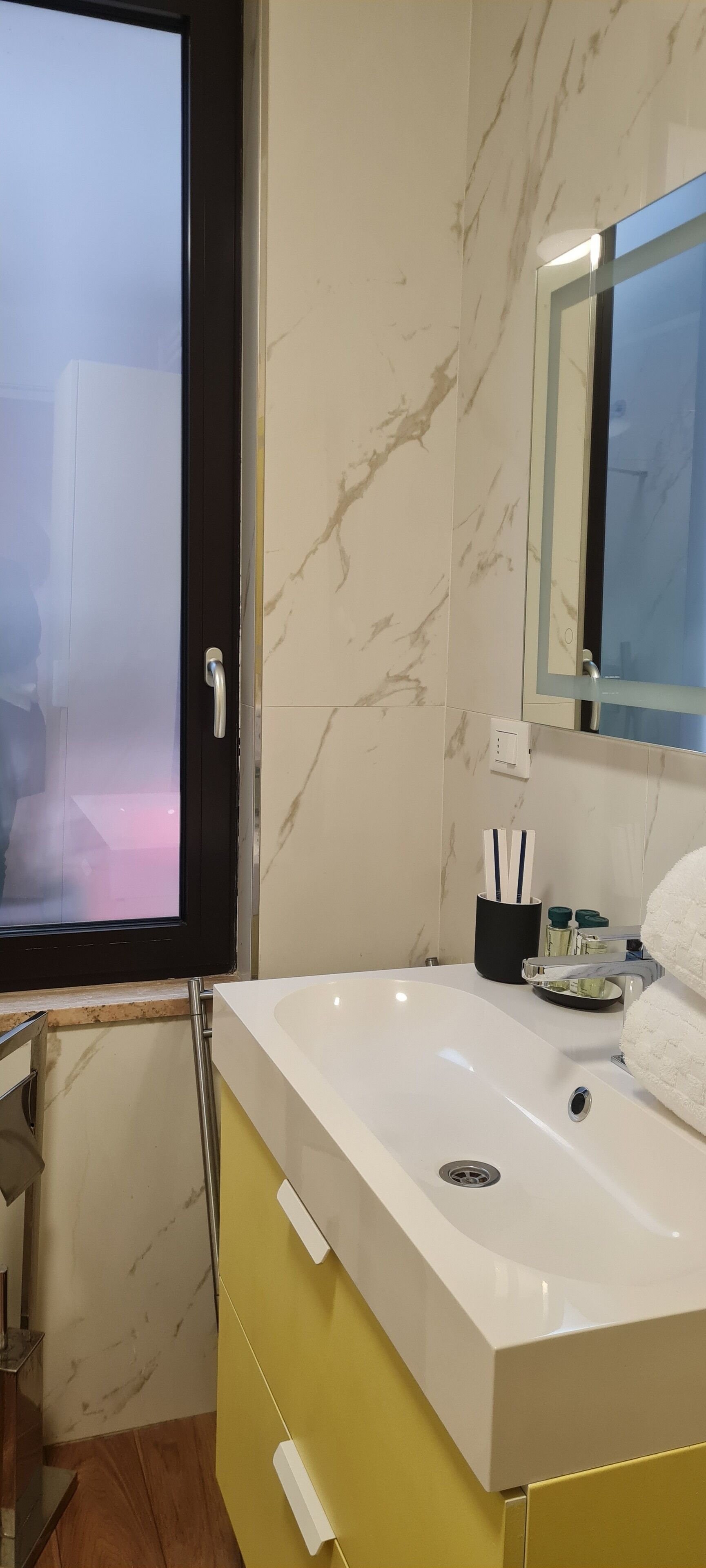 Une salle de bain avec un lavabo blanc, un meuble jaune, un mur en marbre et un grand miroir.