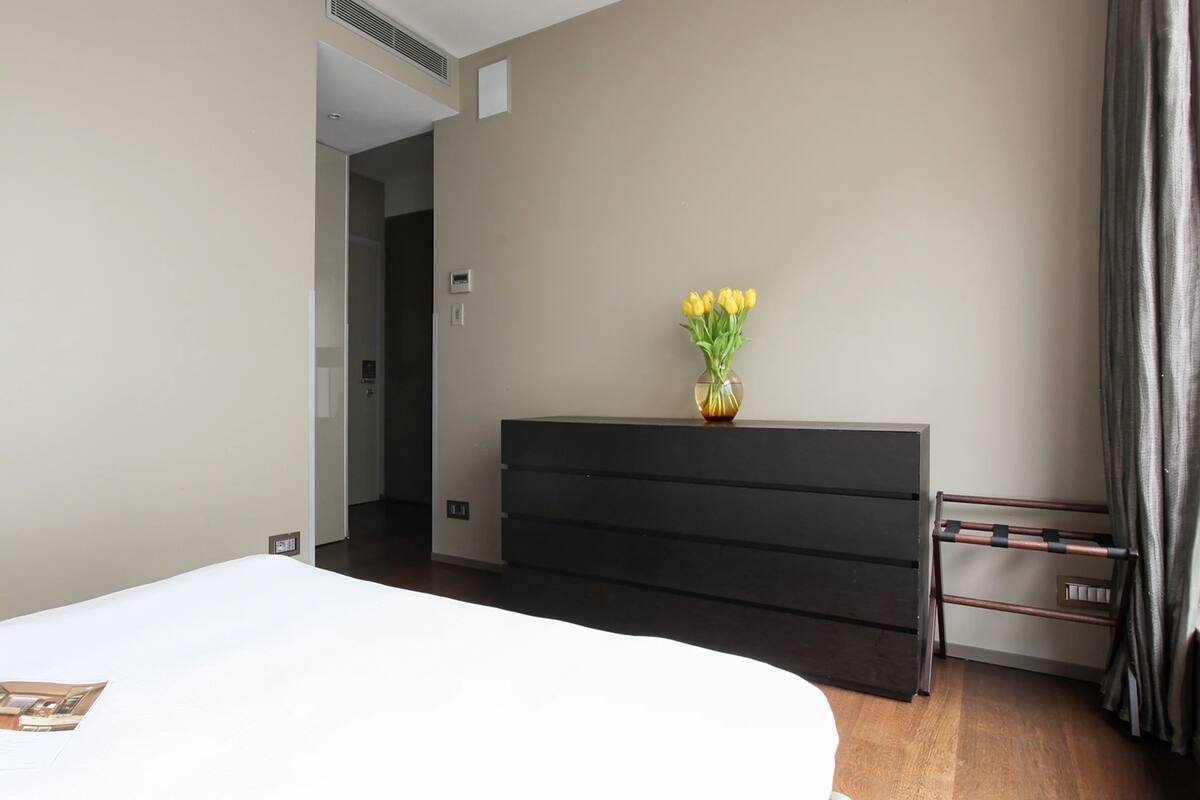 Ein Hotelzimmer mit einem Bett, einer schwarzen Kommode, einem Blumenstrauß mit gelben Blumen und einer Zeitschrift auf dem Bett.