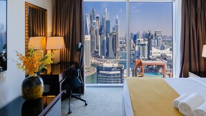 A hotel room with a large window offering a city skyline view, a bed with white linens, a desk with a lamp, and a vase with flowers. - Stylish 1BR unit Address Marina (Dubai)