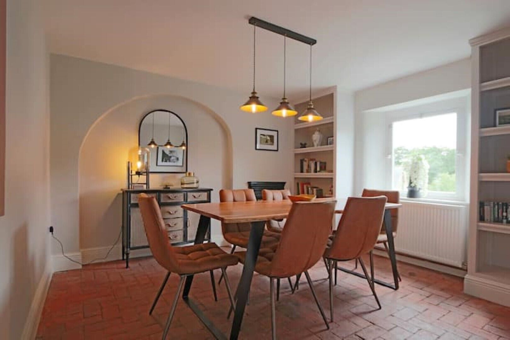 Un coin repas avec un sol en briques, une table en bois et six chaises. On y trouve une bibliothèque remplie de livres et d’objets décoratifs, ainsi qu’une fenêtre donnant sur un paysage verdoyant.
