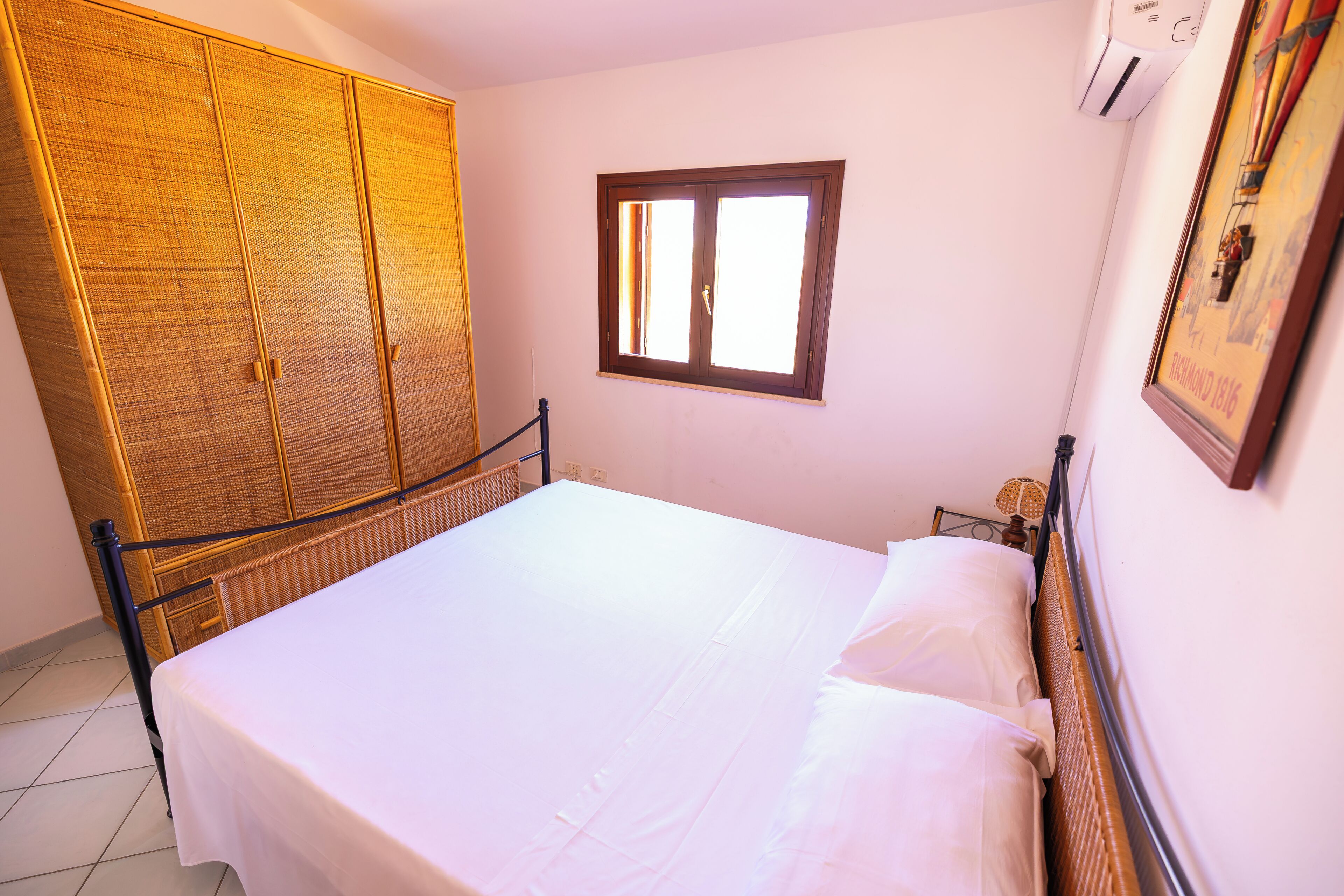 A bed with a white sheet, a wooden wardrobe, a window, and a wall-mounted air conditioner.