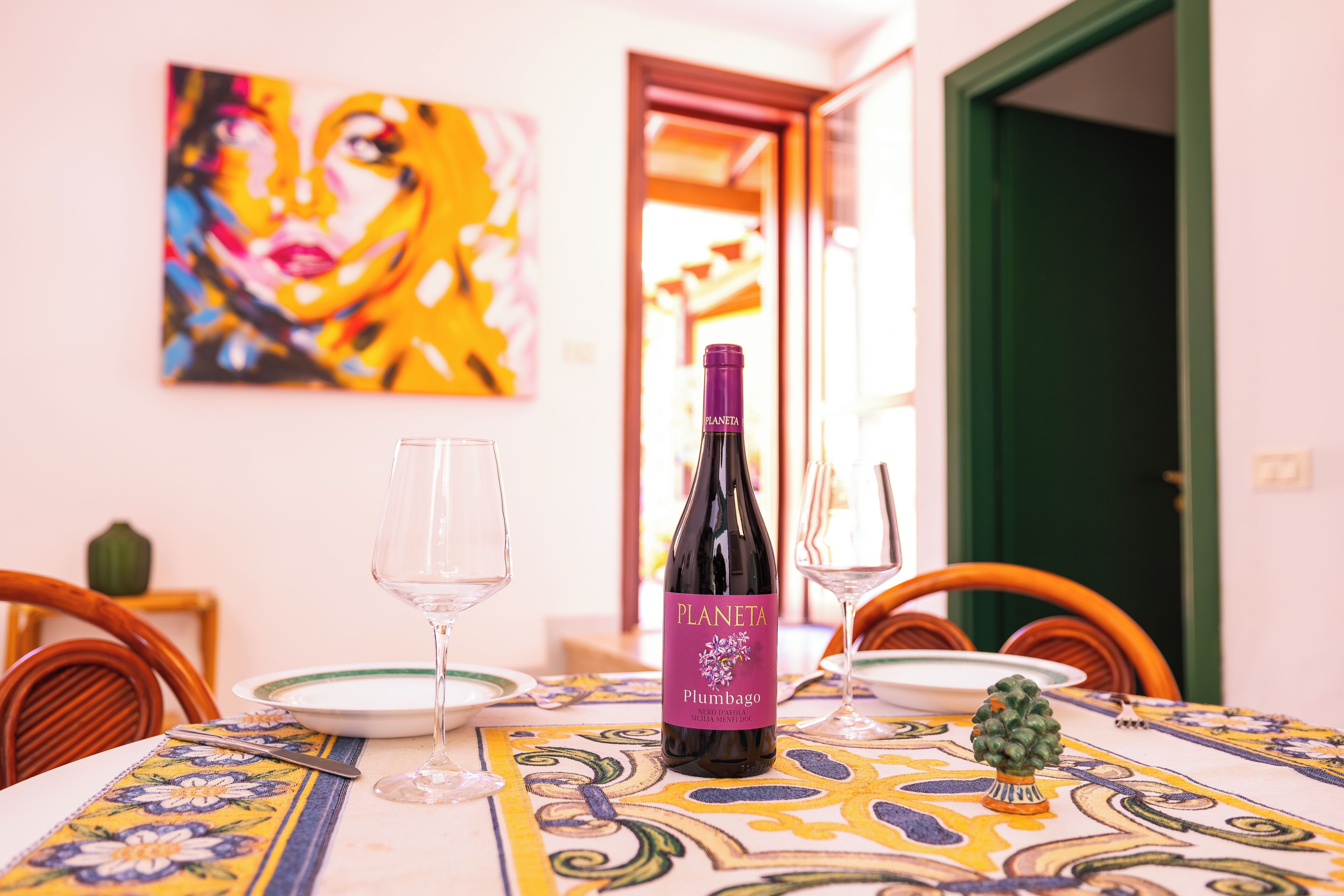 Uma mesa de jantar com uma garrafa de vinho Planeta Plumbago, duas taças de vinho e um pequeno vaso de planta.