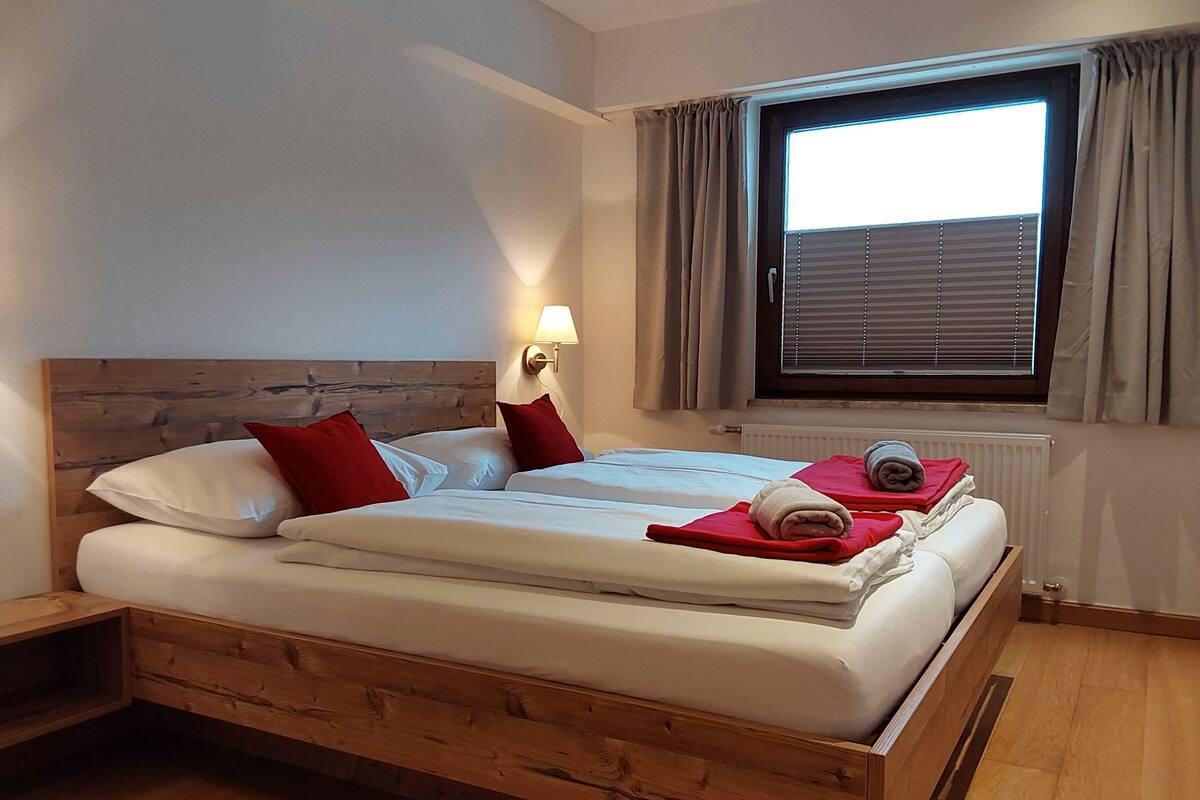 A double bed with wooden headboard, white bedding, red pillows, and a bedside lamp. There is a window with blinds and curtains, and a radiator below.
