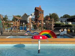 Un espace de jeux pour enfants avec un toboggan, des structures d’escalade et une piscine agrémentée d’une fontaine en forme de champignon.