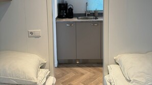 Une chambre de taille modeste, équipée de deux lits, d’une kitchenette et d’une fenêtre avec des stores.