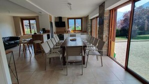 A dining area with a long table and chairs, a kitchen area with a stove and sink, and a large sliding glass door to a garden.