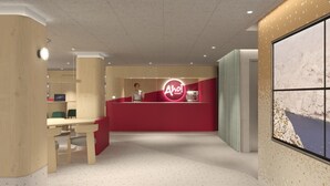 A reception area with a red counter, a woman standing behind it, and a large window with a view of a rocky coastline. - AHOI! Urban Beach Hotel (Palma de Mallorca)