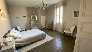 A bedroom with a large bed, a chandelier, a fireplace, and a chair. - Le Mas de la Coquille (Castries)