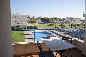 En balkon med et bord og stole, der kigger ud over en swimmingpool og en grøn græsplæne med palmer.
