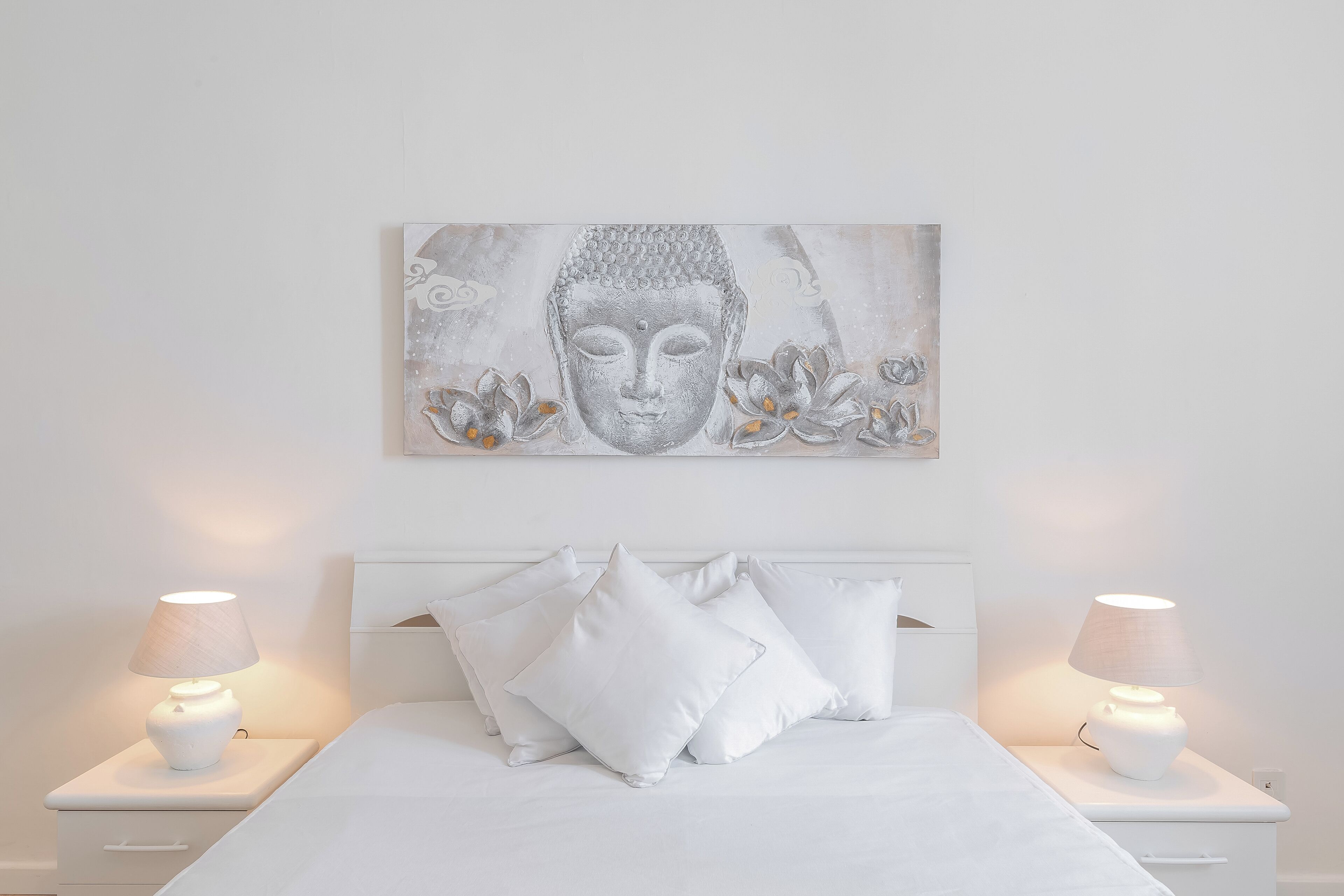 A bedroom with a white bed, two bedside tables with lamps, and a wall-mounted artwork of a Buddha.