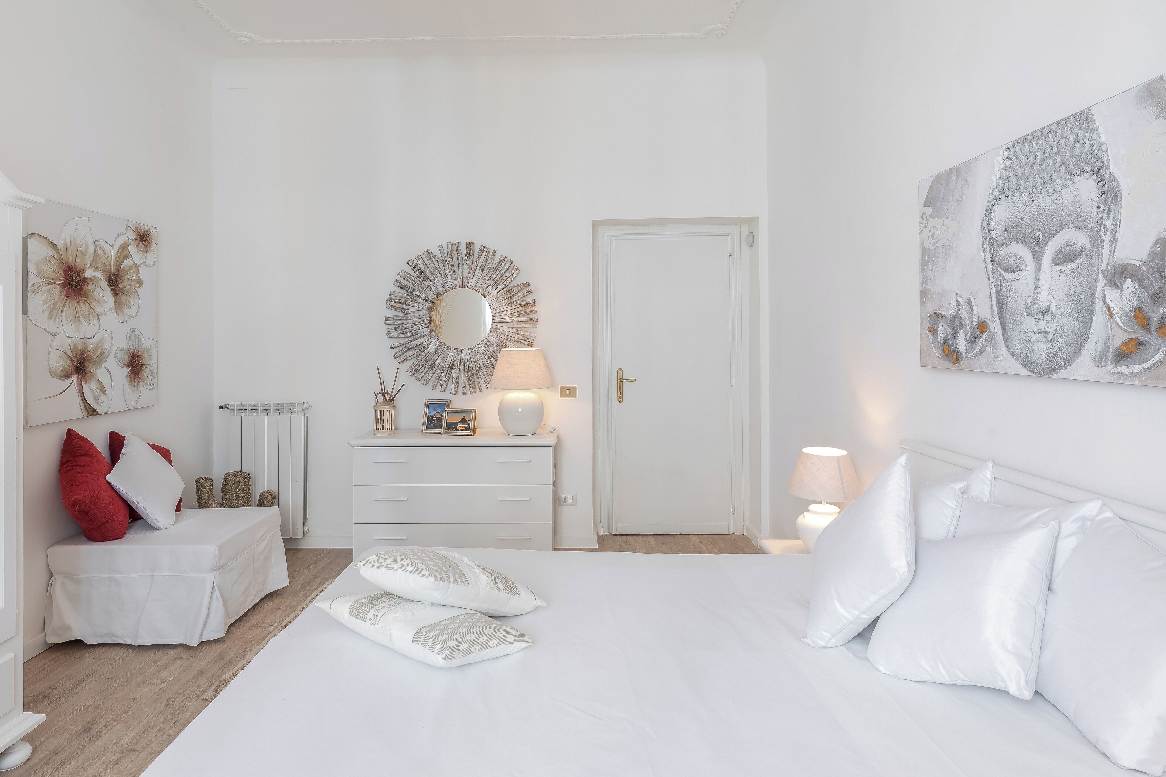 A minimalist bedroom with a white bed, a bedside table with a lamp, a dresser, and a large mirror.