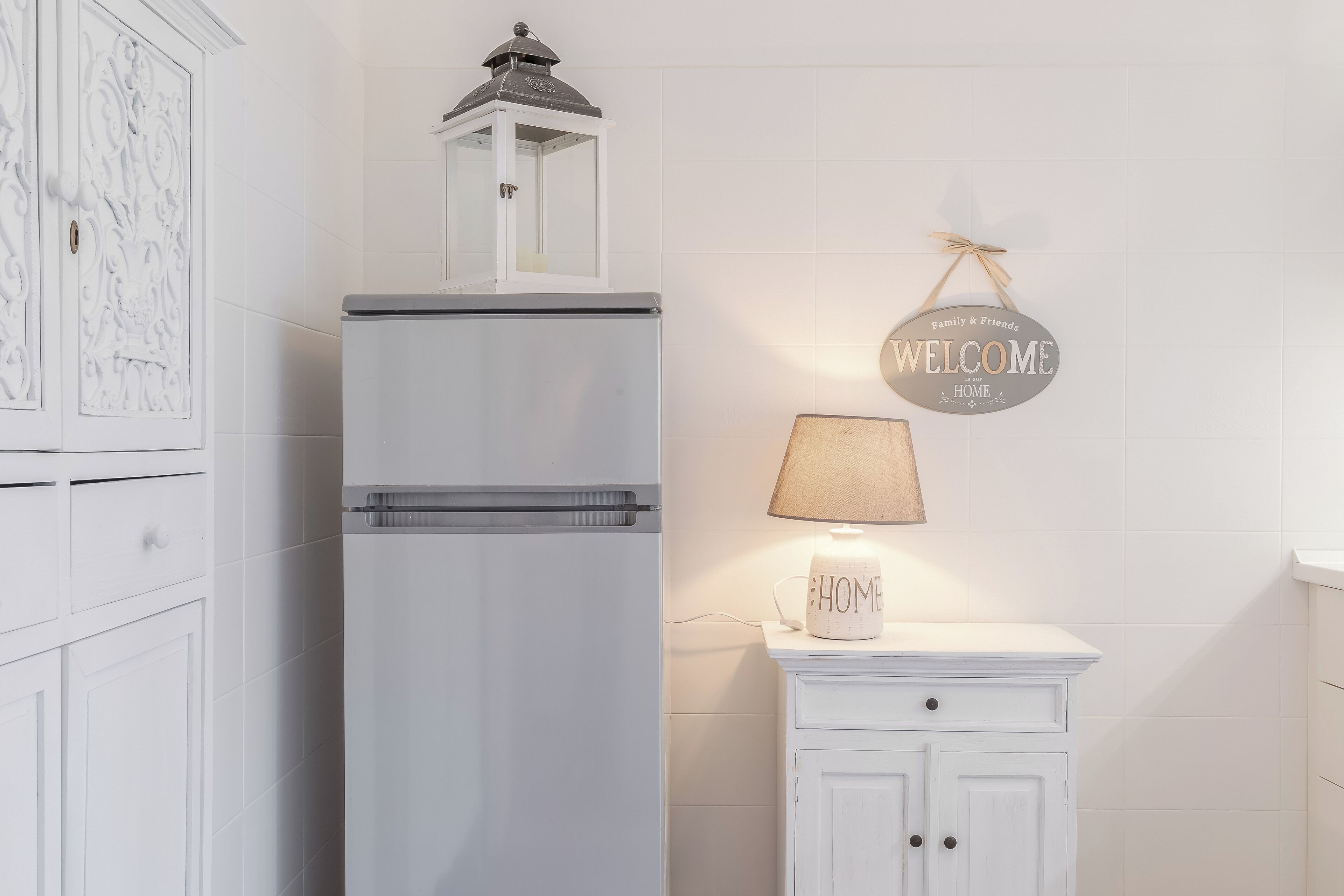 Un réfrigérateur blanc surmonté d’une petite lanterne, un meuble blanc avec une lampe, et une pancarte « BIENVENUE À LA MAISON » accrochée au mur.