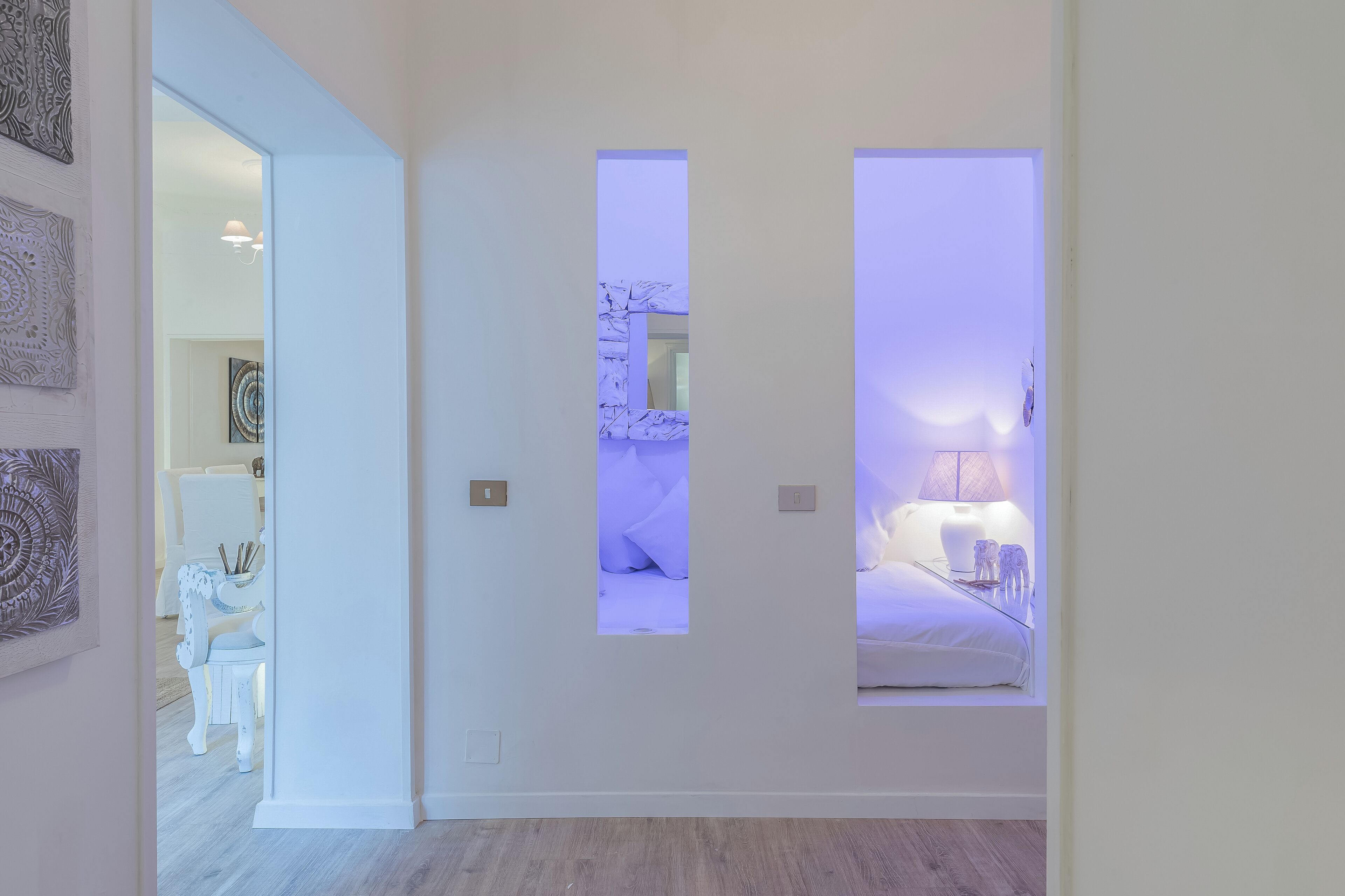 A modern interior with a hallway leading to a bedroom. The room features a bed with a nightstand and lamp, a white chair, and a small table with decorative items.