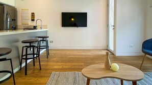 A modern living room with a kitchen area, wooden flooring, a flat-screen TV mounted on the wall, and a blue chair. - w* | Bright 2BR Duplex with Balcony in Barranco (Barranco)