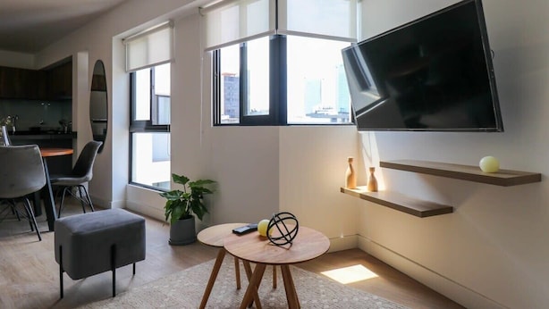 A modern living room with a flat-screen TV mounted on the wall, a round wooden table, and a seating area with a grey ottoman and chairs.