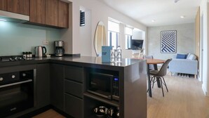 A modern kitchen with dark cabinetry, a built-in oven, and a microwave. There is a dining area with a table and chairs, and a sofa in the background.