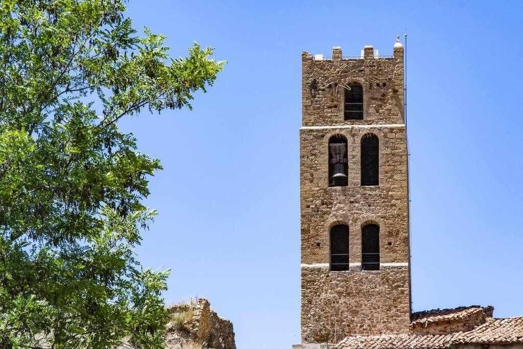 Una torre alta i històrica de pedra amb finestres d'arc i una part superior amb merlets, envoltada d'arbres i sota un cel serè.