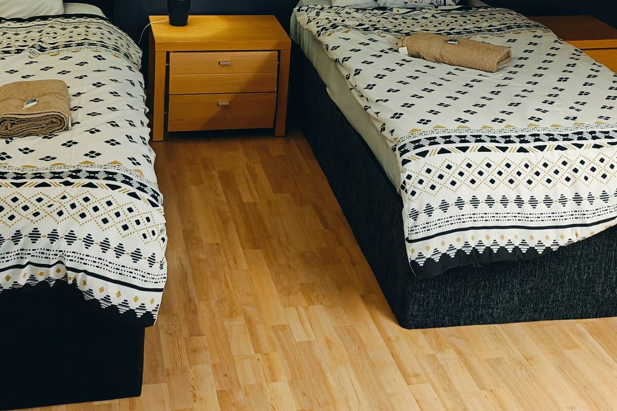 A small, cozy room with two beds, a nightstand with a lamp, and a wooden floor.