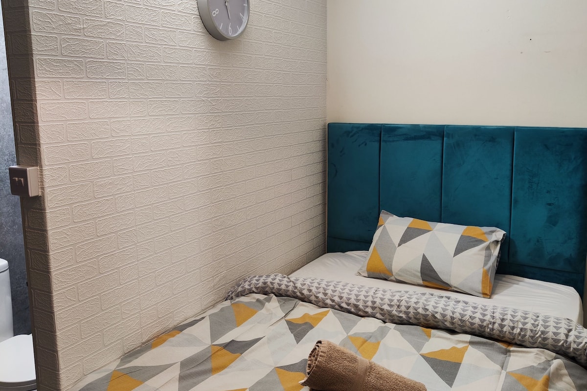 A bedroom with a brick-patterned wall, a clock, a teal headboard, and a bed with patterned bedding.