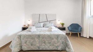 Une chambre moderne avec un grand lit, deux tables de chevet, une chaise bleue et une vue sur l’extérieur.