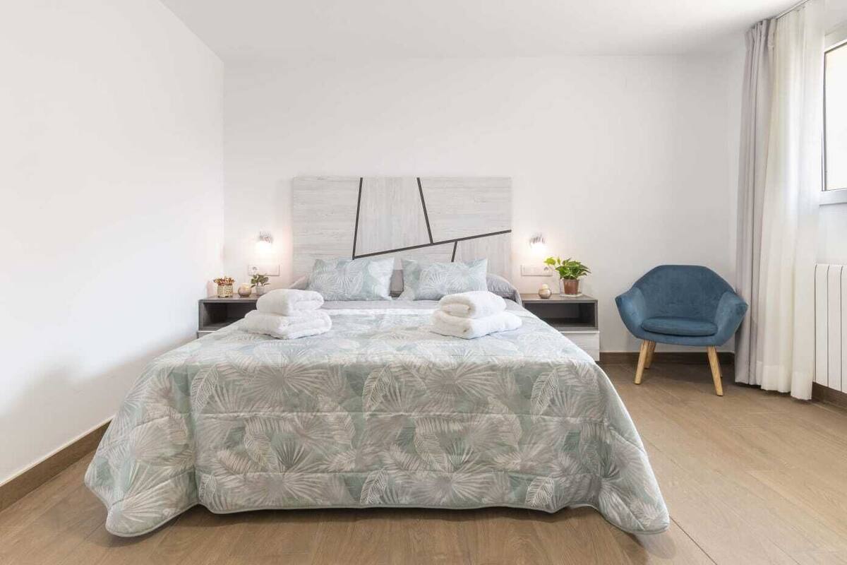 A modern bedroom with a large bed, two bedside tables, a blue chair, and a view of the outside.