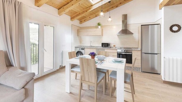 Une cuisine moderne avec des poutres en bois, un coin repas avec une table et des chaises, et une vue à travers les portes vitrées.