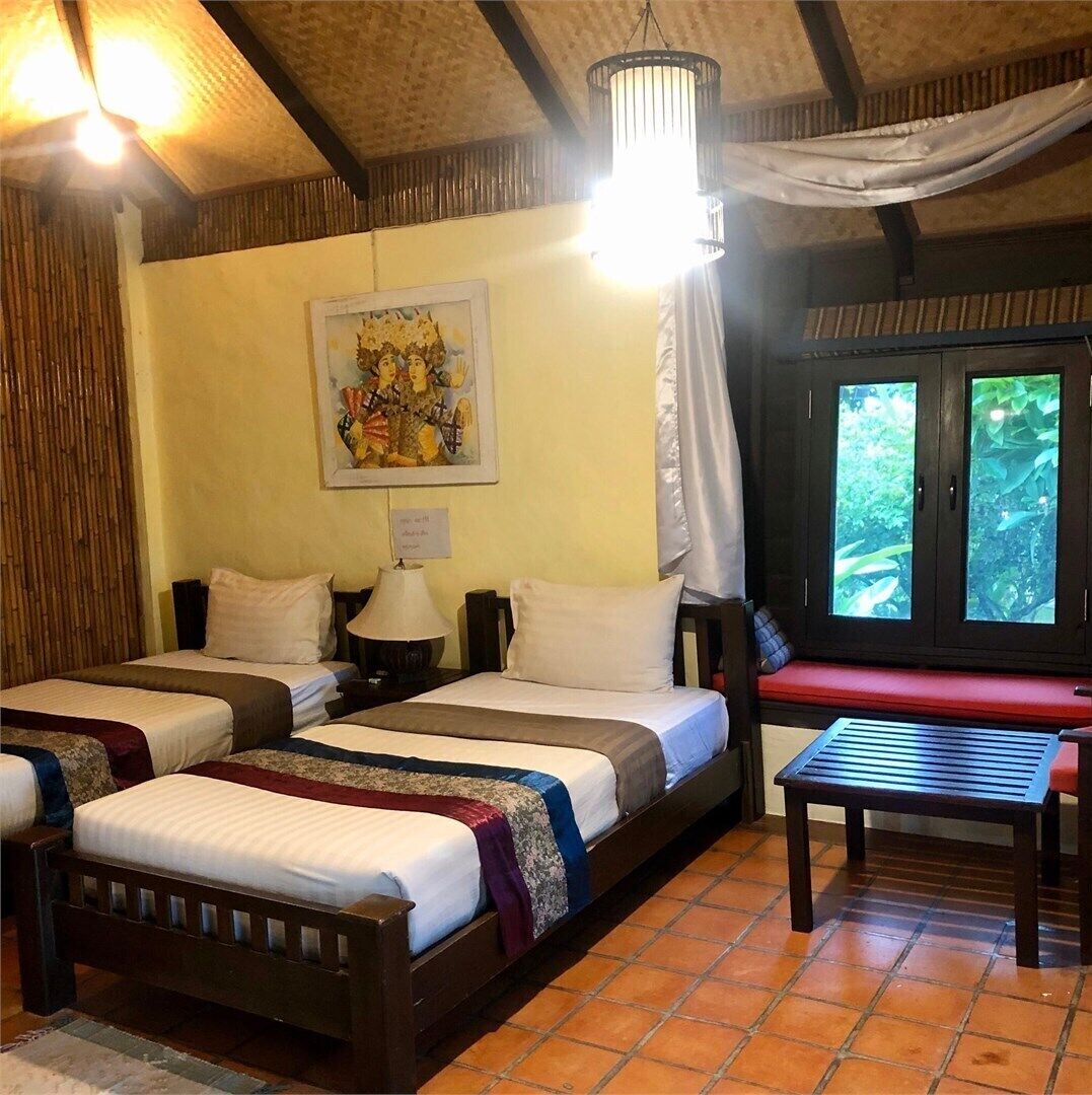 A room with two beds, a bench, a table, a painting, and a window with curtains.