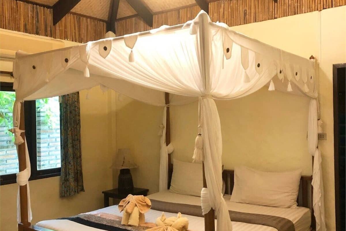 A four-poster canopy bed with white linens and a wooden frame, a chandelier, and a window with curtains.