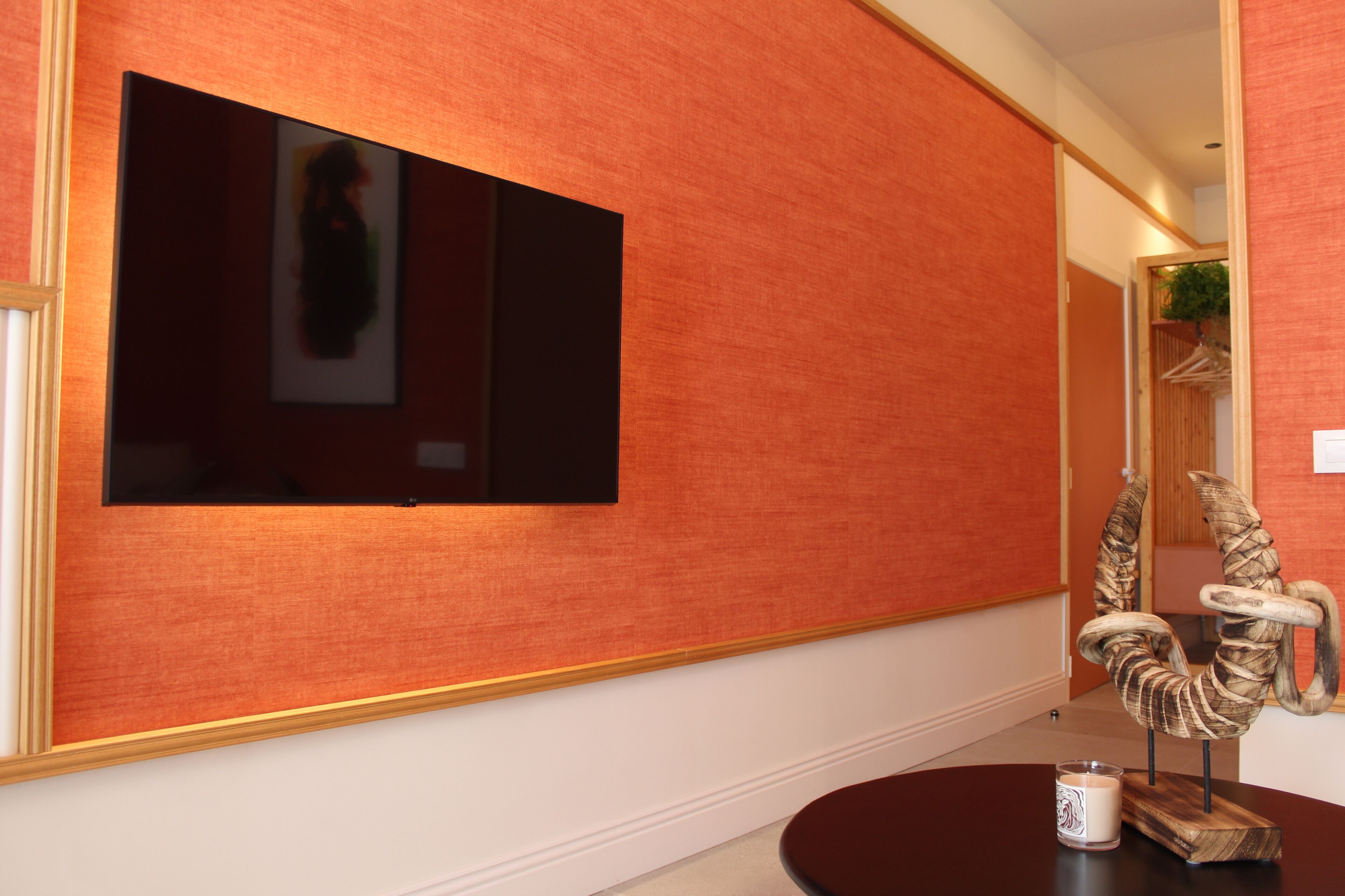 A room with a large flat-screen TV mounted on a textured orange wall, a wooden chair with a unique design, and a small table with a candle.